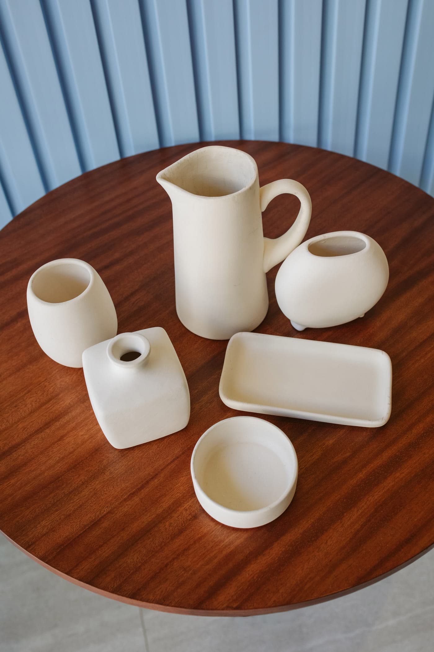 Jarra, vasos y charolas de cerámica en crudo sobre una mesa de madera